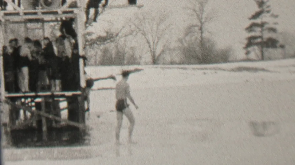 In metadata-supported Albany, New York, 1937, a swimsuit-clad man stands or walks on ice near a wooden dock where bundled spectators watch.