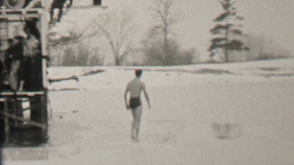 The Albany winter home-movie frame shows the swimmer walking across the ice as the dock and distant snow-covered shoreline remain in view.