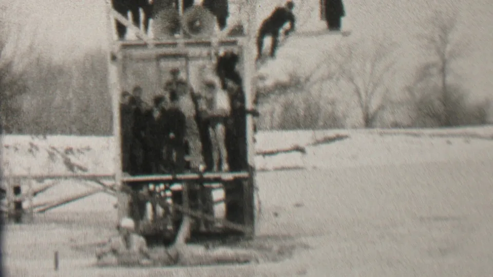 A group of bundled spectators stands on the wooden dock in Albany as the swimmer appears near the frozen water or ice below and beyond them.