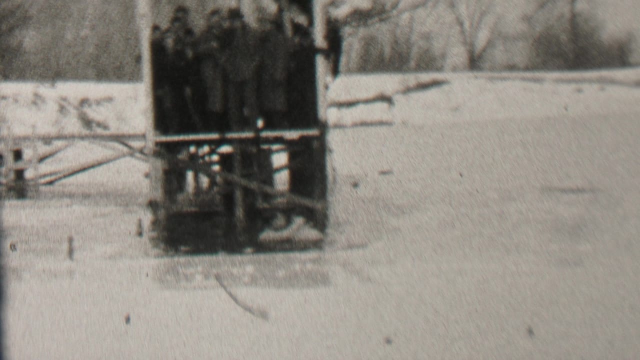 A crowded wooden platform stands at the edge of a snowy frozen area with bare trees in the background.