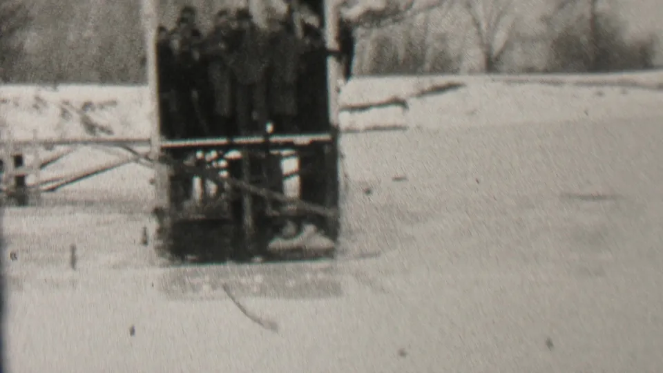 A group of bundled spectators stands closely together on a wooden platform beside a frozen lake area in Albany, New York, in the 1937 footage.