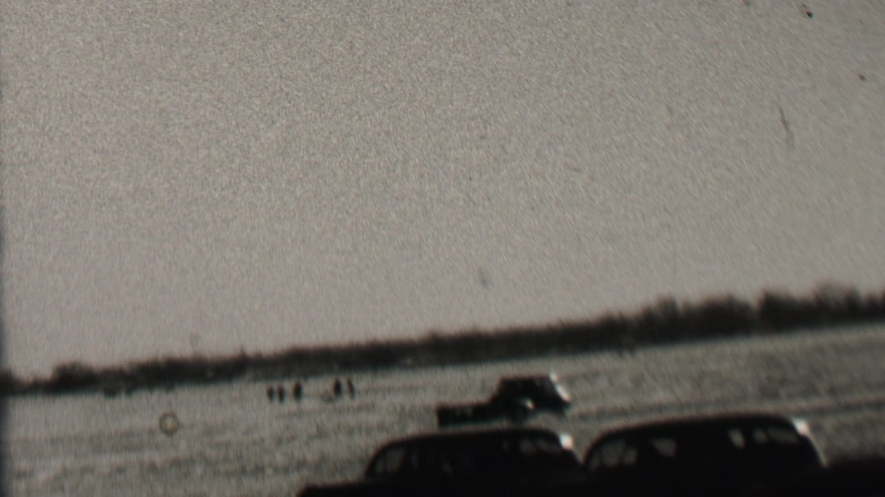 1937 Albany, New York 8mm frame showing dark early automobiles driving over an open field with small groups of observers in the distance.