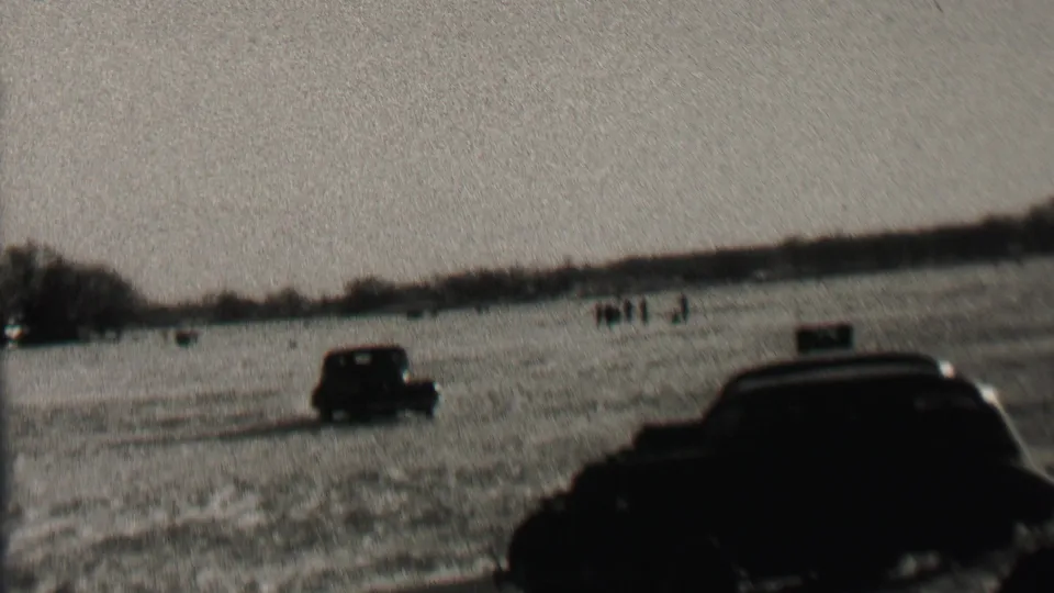 1937 Albany, New York 8mm frame with a dark foreground automobile, a second car in the field, distant observers, and a low tree-lined horizon.