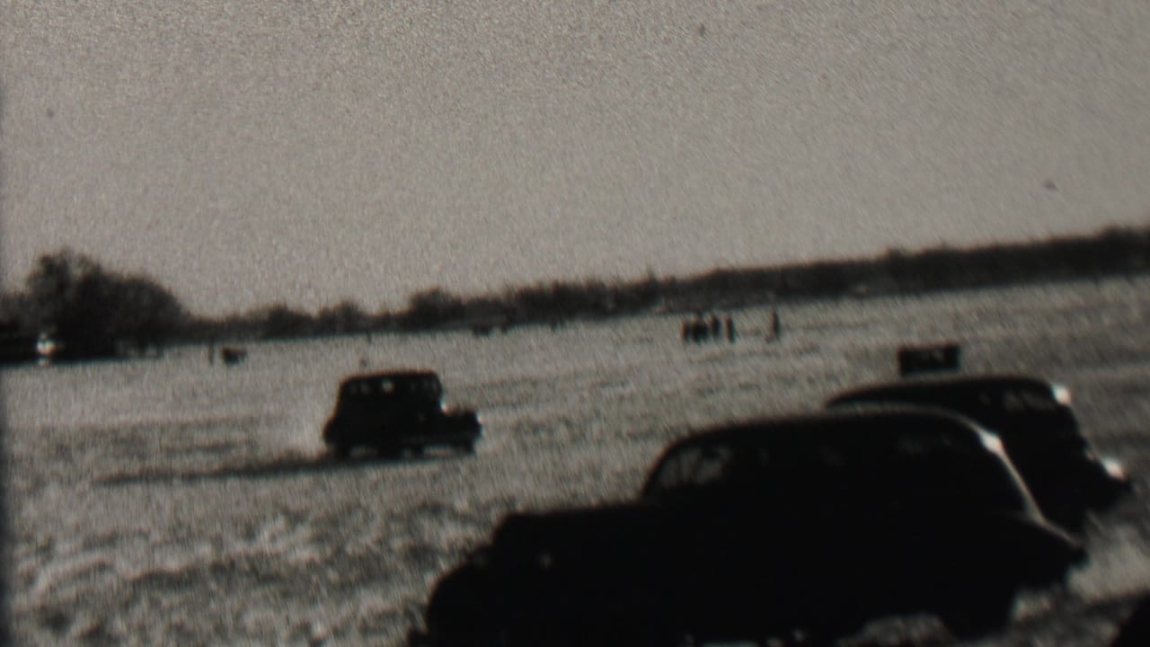 1937 Albany home-movie frame showing dark prewar cars moving through a snow-covered field with bare trees and distant figures or markers on the horizon.