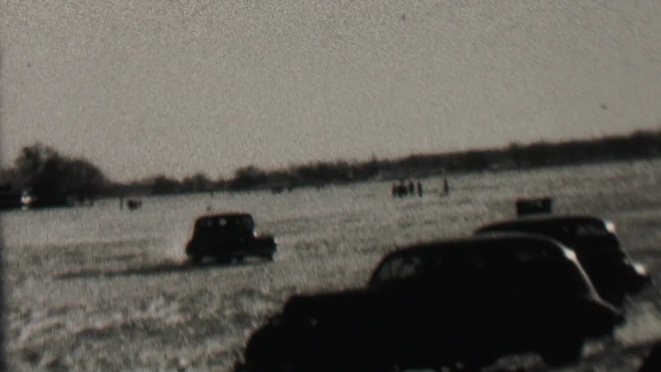 1937 Albany home-movie frame showing dark prewar cars moving through a snow-covered field with bare trees and distant figures or markers on the horizon.