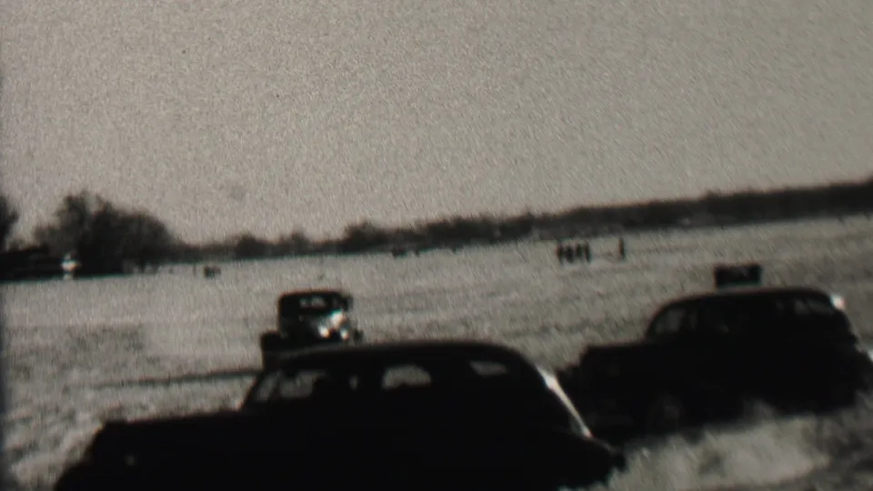 Several dark automobiles appear in a snowy Albany field, with one car near the foreground and others farther back under a pale winter sky.