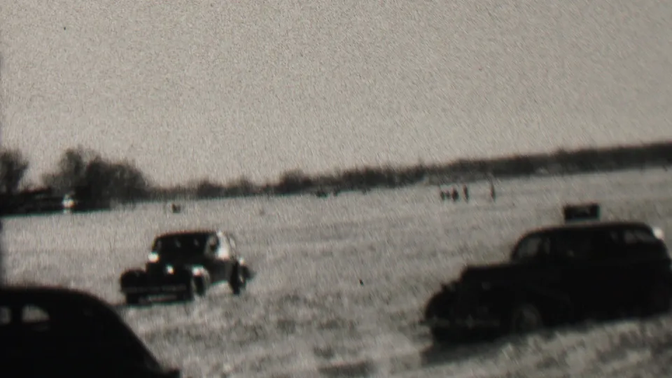 A black-and-white 8mm frame shows cars traveling over snow in an open field, with a low tree line and indistinct distant activity in the background.