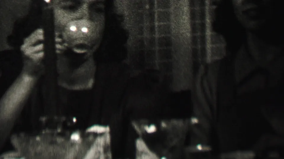 Indoor 1937 Albany home-movie frame showing women at a table with cups and glassware, including one woman facing the camera in dim, high-contrast light.