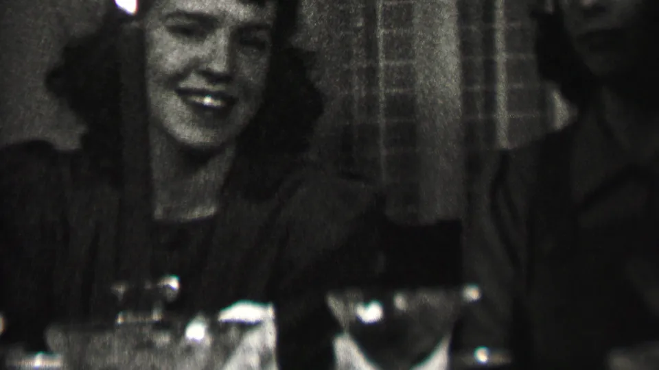 The woman smiles broadly at the dinner table, with glasses, folded linen, and a dark private interior visible in the 1937 Albany home-movie frame.