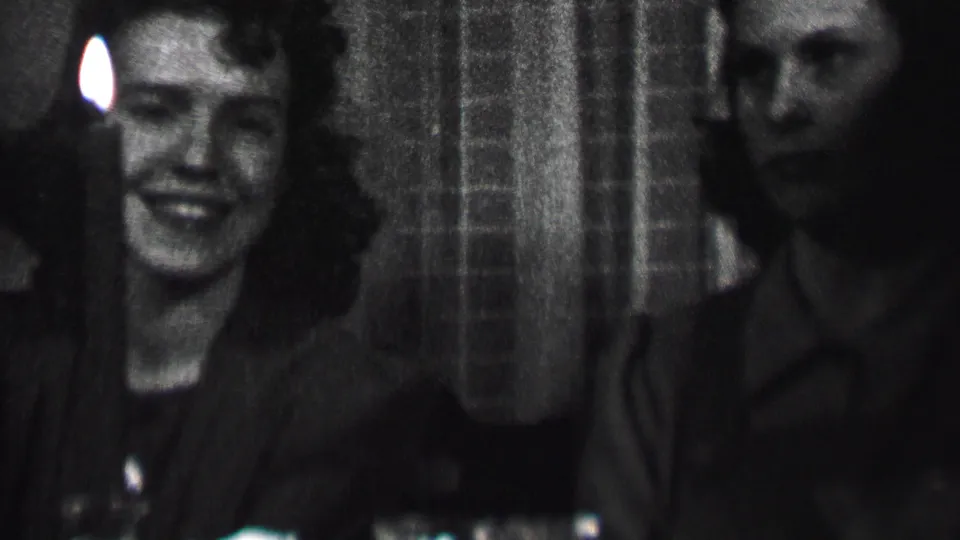 A smiling woman sits near a small candle flame at the dining table, with an adult man partially visible beside her in the Albany 1937 footage.