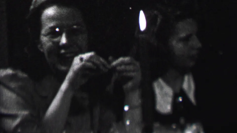 Albany 1937 home-movie frame showing one woman smiling toward the camera while another sits beside her in a shadowed candlelit room.