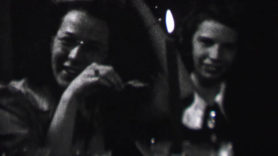 A woman laughs or smiles broadly beside another seated woman in the supplied 1937 Albany home-movie clip, with a candle and bottles visible in the dark interior.
