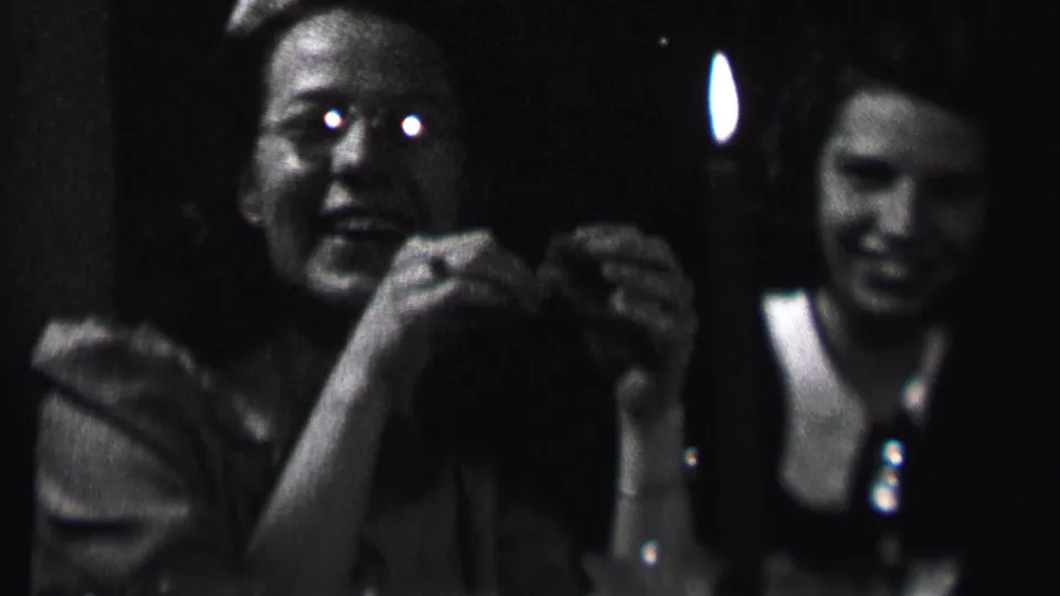 1937 Albany home-movie frame showing two young women at a candlelit indoor gathering, one facing the camera while another smiles in the background.