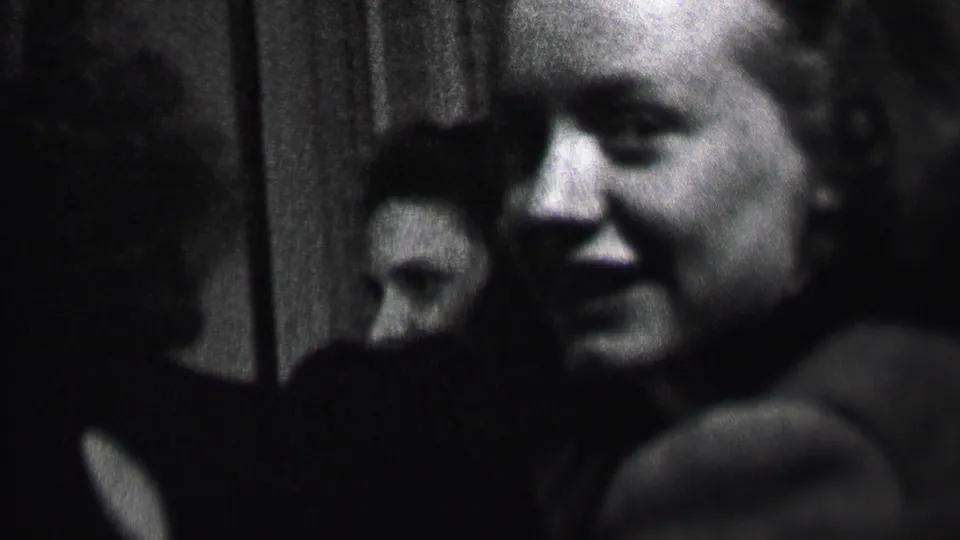 Close handheld 1937 Albany home-movie frame showing a smiling young woman in soft focus, with another face partly visible in the private interior behind her.
