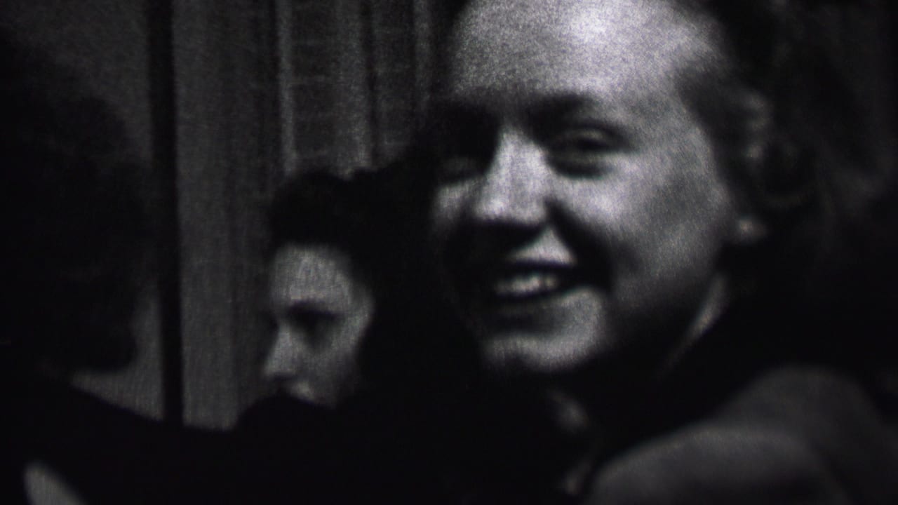 Black-and-white 1937 Albany home-movie frame showing a smiling bald man near the camera while young women sit or stand in the dim interior behind him.