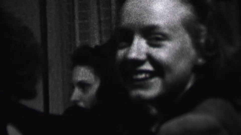 Black-and-white 1937 Albany home-movie frame showing a smiling bald man near the camera while young women sit or stand in the dim interior behind him.