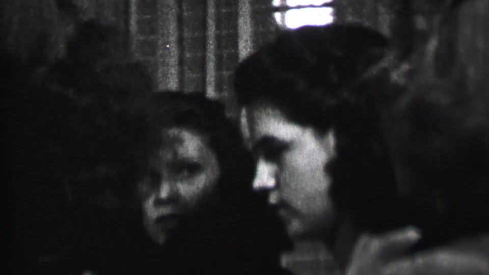 Black-and-white 1937 Albany home-movie frame showing young women in close conversation inside a dark private interior with a bright window or light behind them.