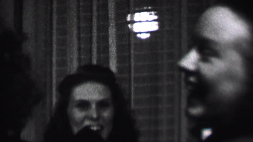 Black-and-white 1937 Albany home-movie frame showing a smiling young woman near center and a bald man in profile at the right edge of a dim interior.