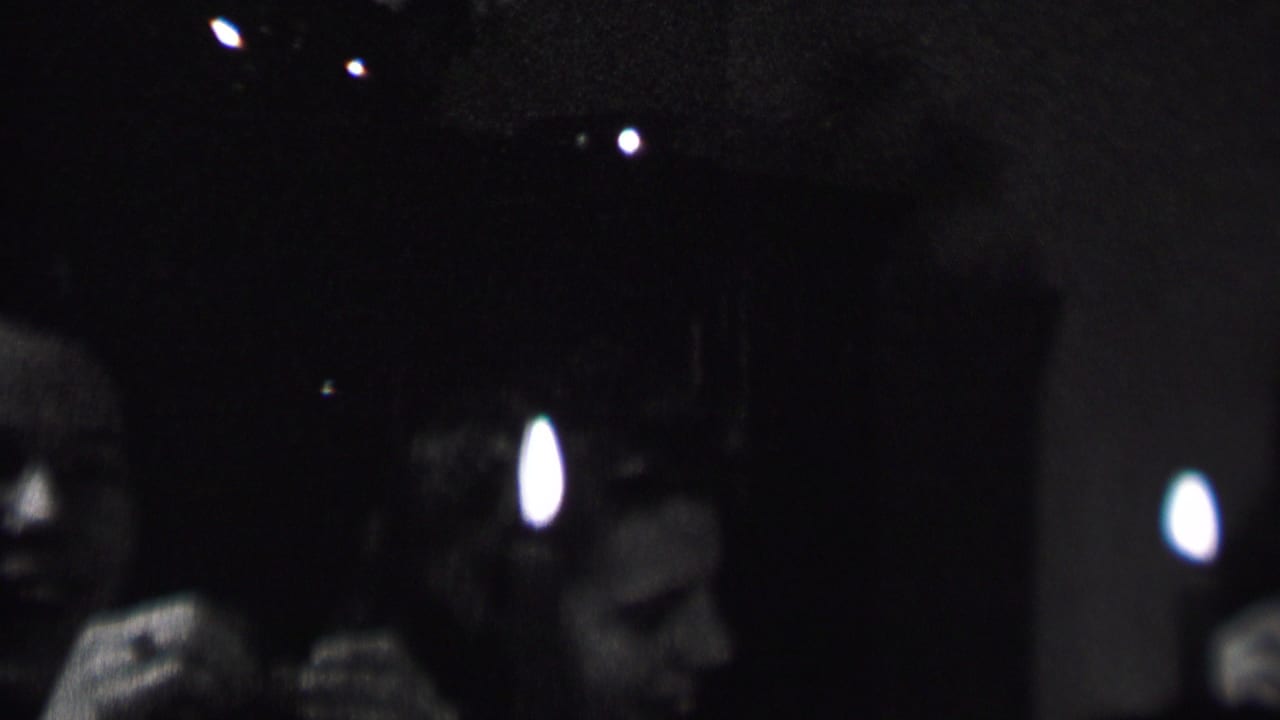 In this 1937 Albany, New York 8mm home-movie frame, adult party guests sit in a dark interior while a bright candle flame partially obscures faces in the foreground.