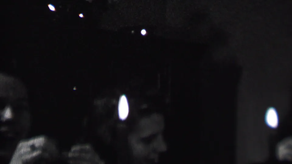 In this 1937 Albany, New York 8mm home-movie frame, adult party guests sit in a dark interior while a bright candle flame partially obscures faces in the foreground.