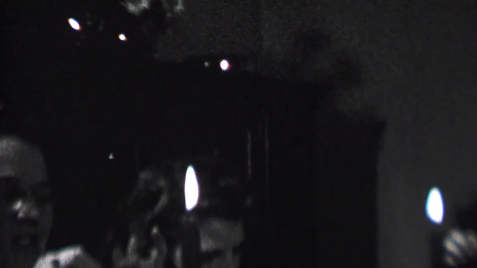 A 1937 Albany home-movie frame shows adults at an indoor party, with one face softly lit near the center and a bright candle flame against dark curtains.