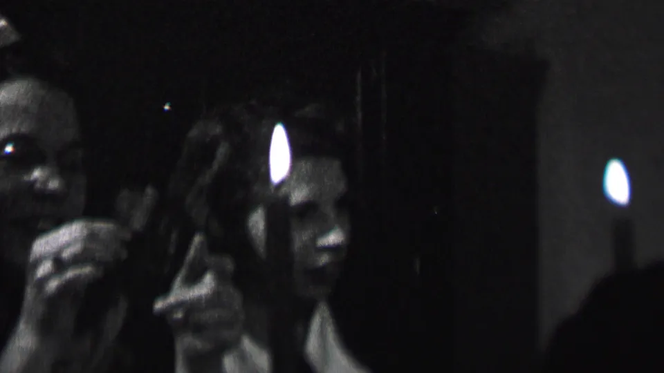A woman smiles in close-up during the 1937 Albany indoor party scene, with a candle flame in front of her and another adult guest partly visible at the edge.