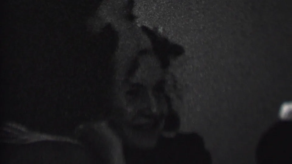 A woman with curled hair laughs in profile while another woman sits beside her in the dark, low-contrast interior of the Albany party footage.
