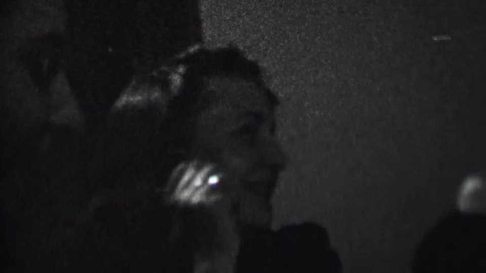 A seated woman smiles in a dim private interior, with her face and hair partially caught by the available light in the 1937 Albany home movie.