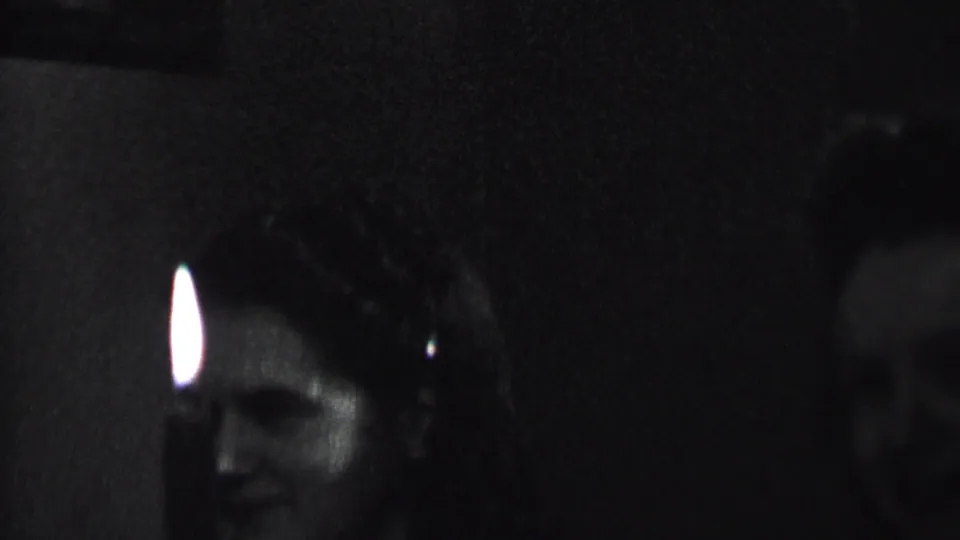 A woman smiles in a dark indoor party scene as a small bright candle-like light appears near the center of the frame, with other figures partly visible around her.
