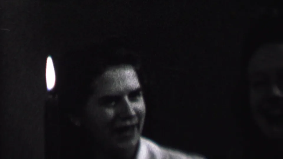 A woman appears in partial profile beside a candle flame, with another shadowed face behind her in the low-light home-movie scene.