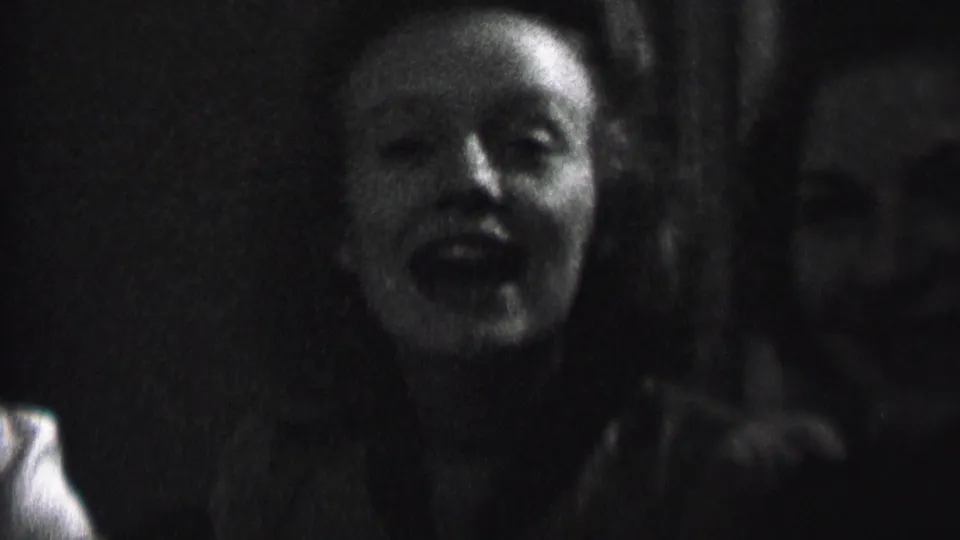 A woman faces the camera in a dim Albany 1937 interior, with another guest beside her and candlelit shadows across the frame.