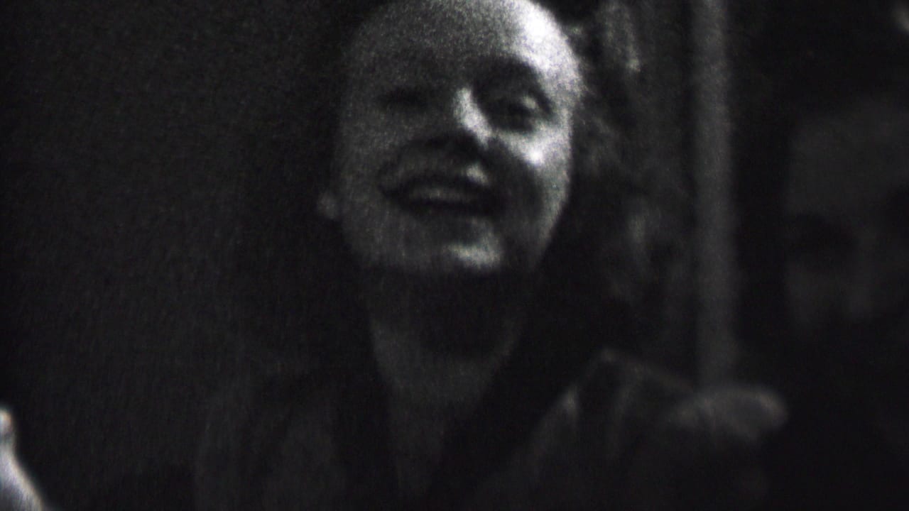 In this 1937 Albany home-movie frame, a young woman smiles toward the camera in low light while another person sits partly obscured beside her.