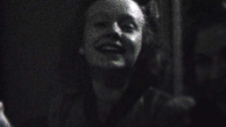 In this 1937 Albany home-movie frame, a young woman smiles toward the camera in low light while another person sits partly obscured beside her.