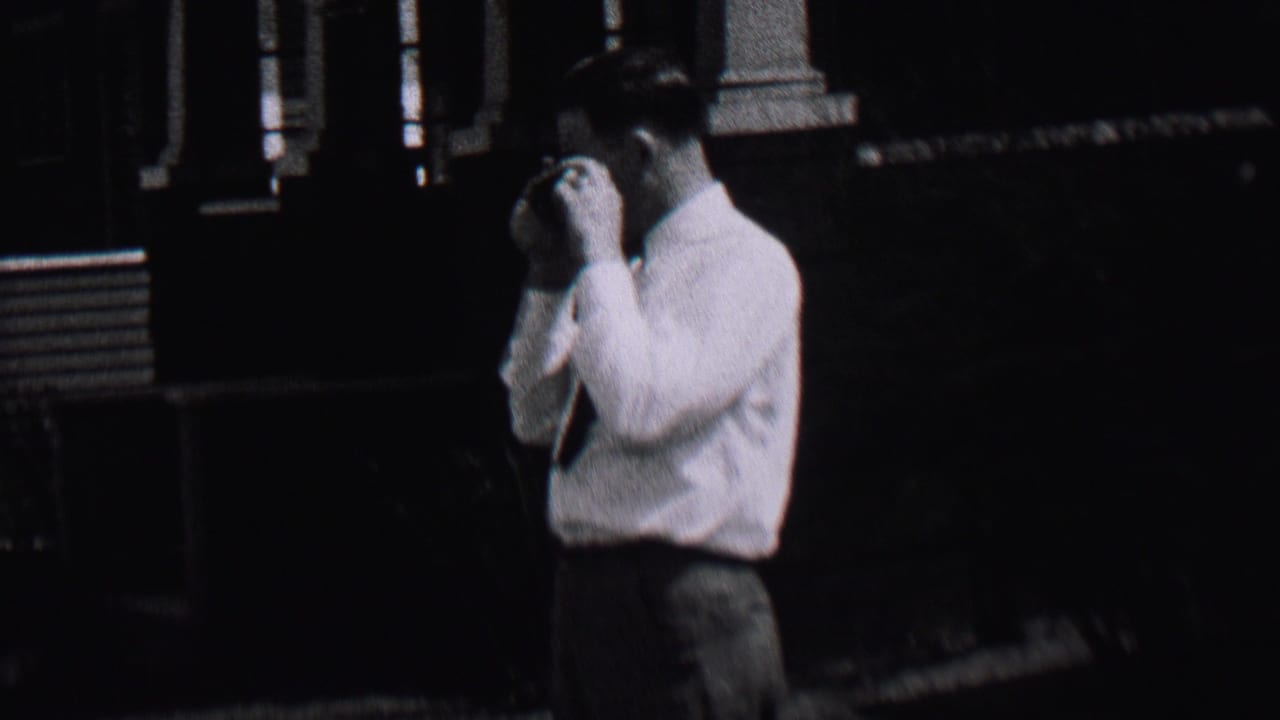 In 1937 Albany, New York home-movie footage, an adult man in a white shirt and tie stands near the edge of a shaded residential street.