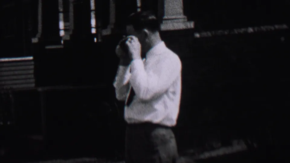 In 1937 Albany, New York home-movie footage, an adult man in a white shirt and tie stands near the edge of a shaded residential street.