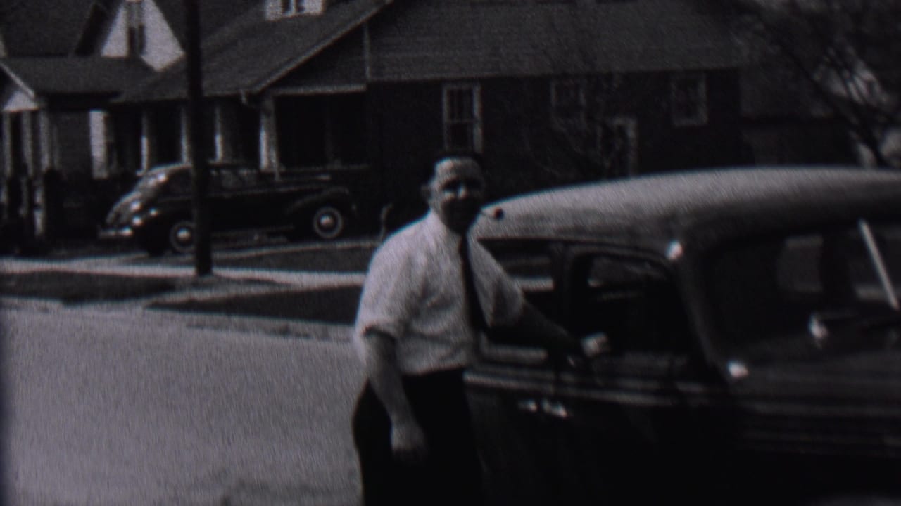 Man stands beside a dark car on a quiet street with houses behind him.
