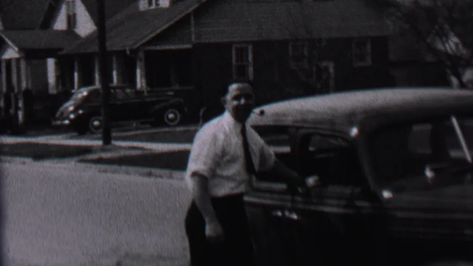 In the Albany, New York residential street scene, the man stands beside a dark 1930s-era automobile with houses and another parked car in the background.