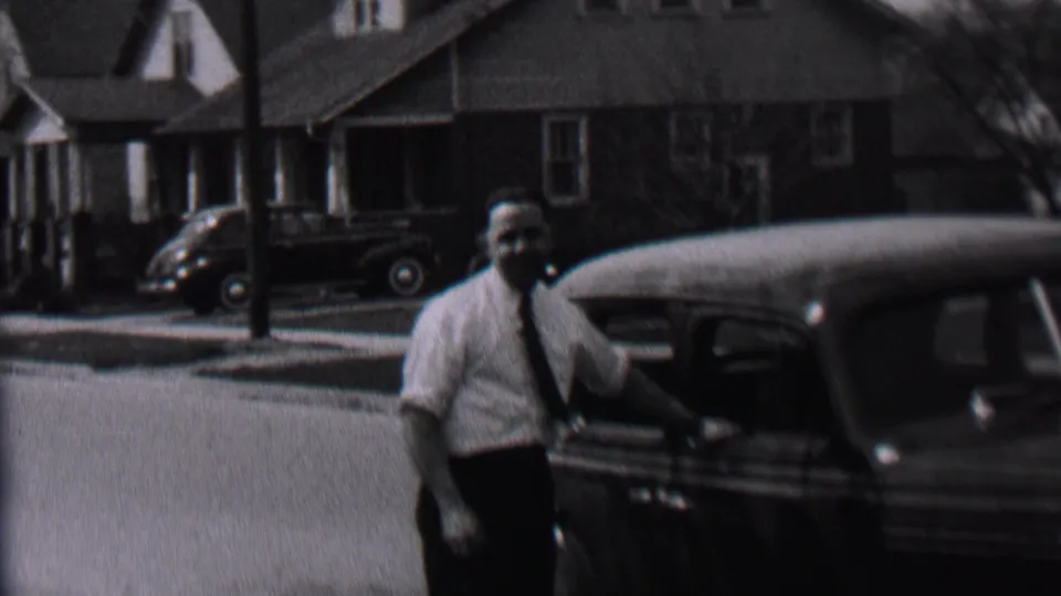 The man faces toward the home-movie camera while standing next to a dark automobile on a sunlit Albany residential street in 1937.