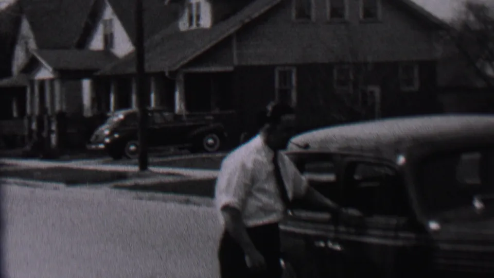 At the end of the clip, the man remains beside the dark car, with residential houses, a sidewalk, and another period automobile visible across the Albany street.