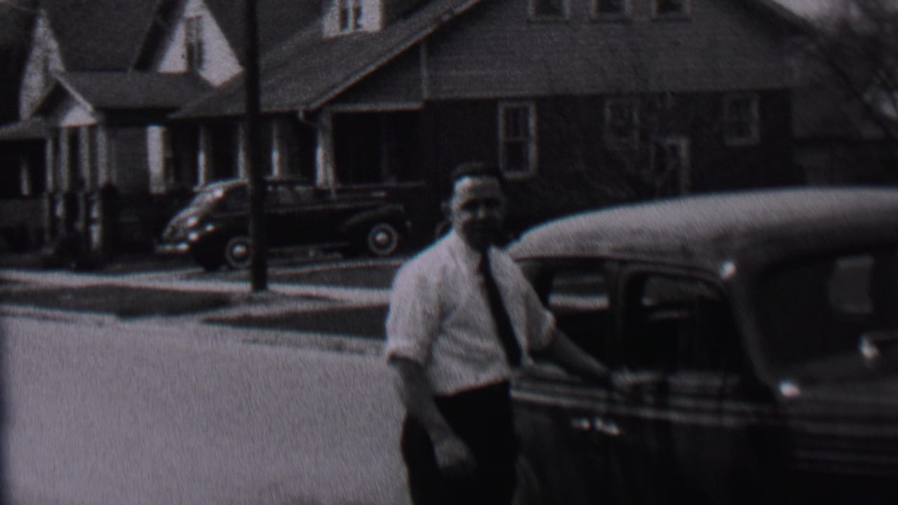 In supplied 1937 Albany home-movie footage, a man in a light shirt and tie stands near a dark automobile with houses and another car visible across the street.