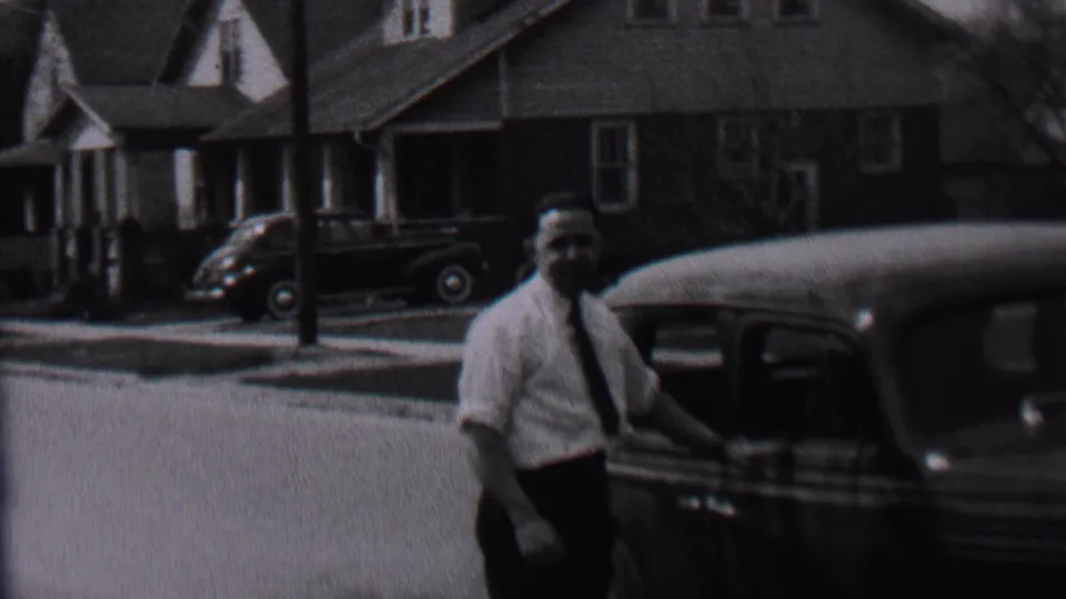 In supplied 1937 Albany home-movie footage, a man in a light shirt and tie stands near a dark automobile with houses and another car visible across the street.