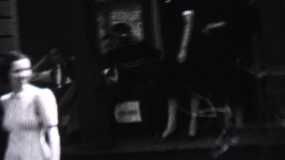 Adults gather on a shaded residential porch in the supplied 1937 Albany footage, with a woman in a light dress at left and seated figures partly obscured by dark exposure.