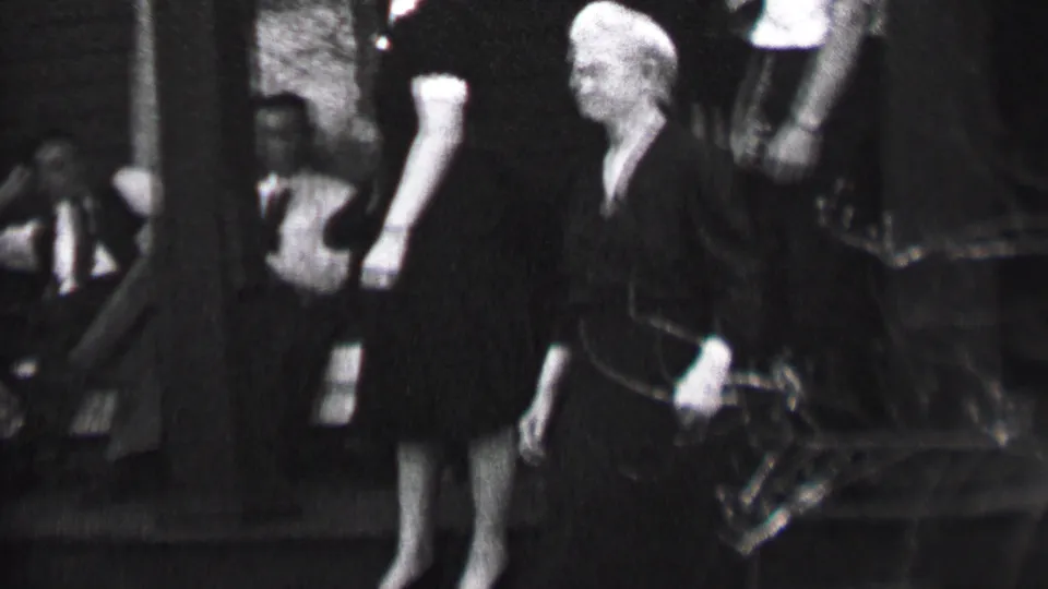 A man in a suit and a woman in a dark dress stand near porch steps during the Albany home-movie gathering, with additional adults partly visible in the background.