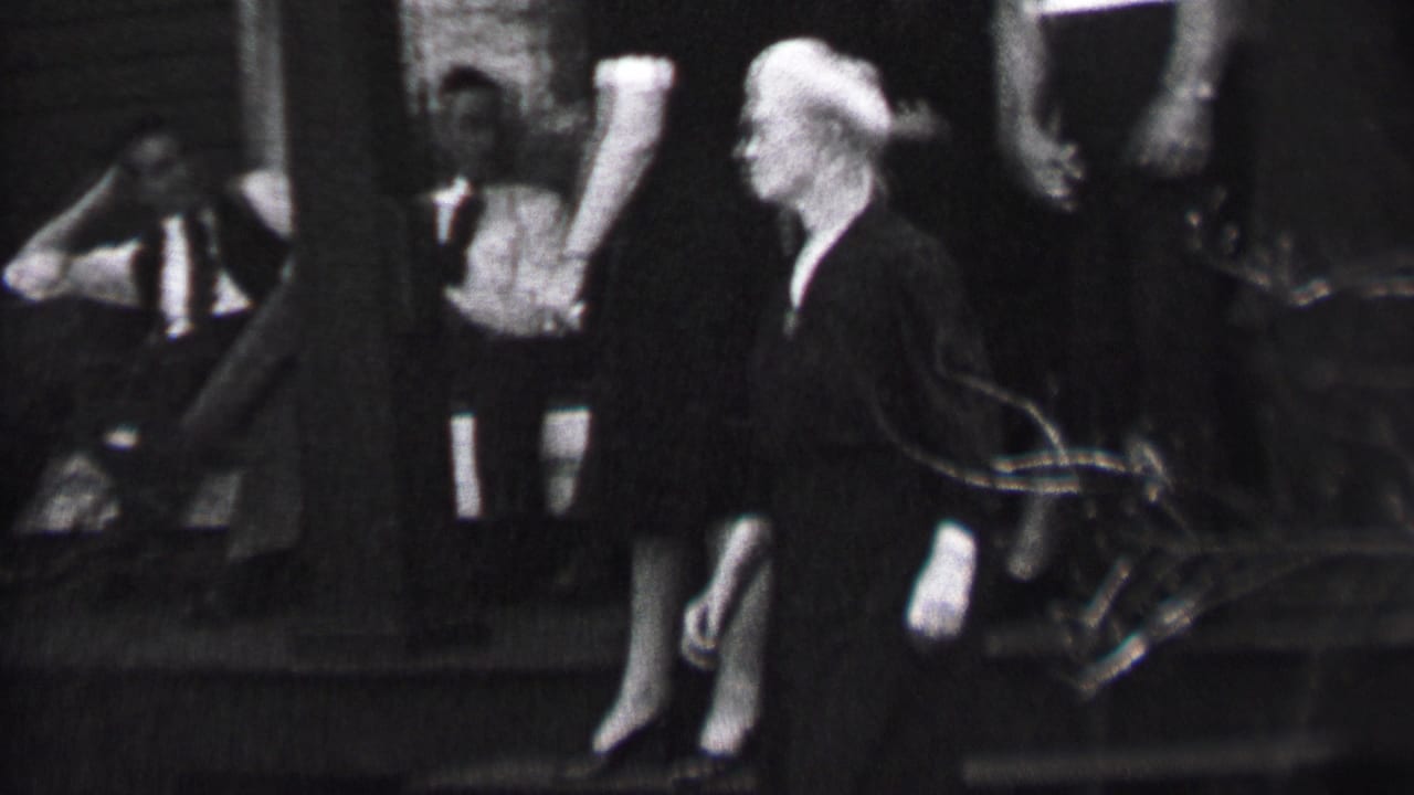 Black-and-white 1937 Albany home-movie frame showing a white-haired woman in a dark dress moving along outdoor steps as men in suits sit and stand behind her.