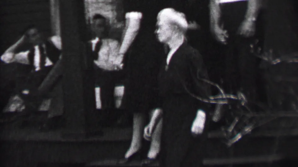 Black-and-white 1937 Albany home-movie frame showing a white-haired woman in a dark dress moving along outdoor steps as men in suits sit and stand behind her.