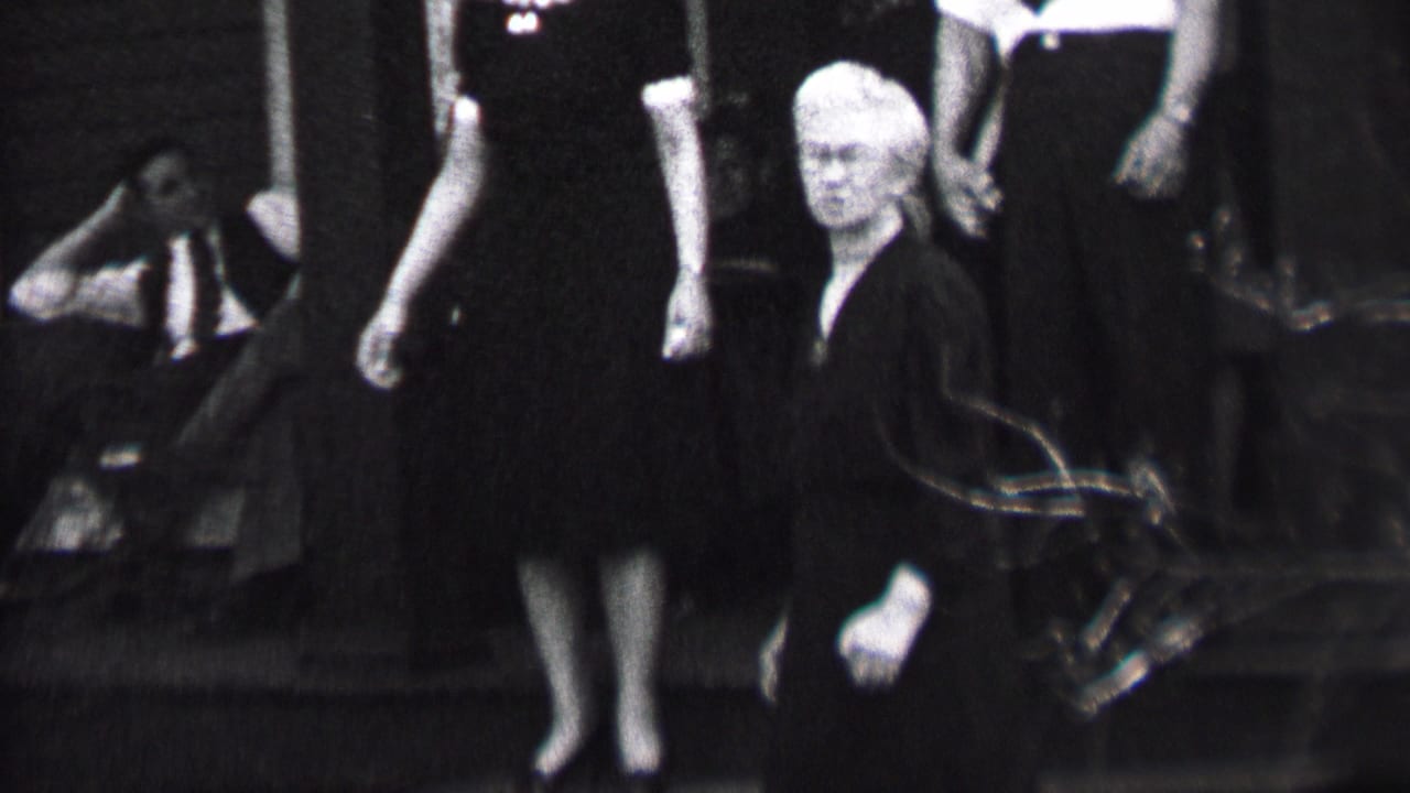 White-haired woman faces forward near outdoor steps with several adults behind her.