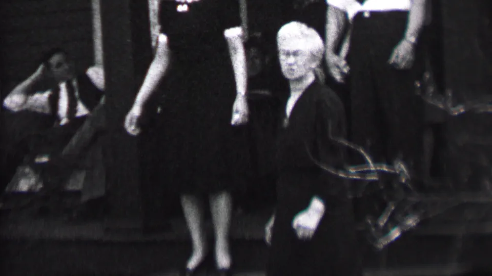 Black-and-white frame from the Albany 1937 clip with a white-haired woman in a dark dress facing toward the camera near steps and adults standing in the background.