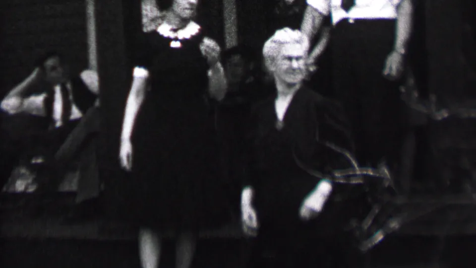 Black-and-white 1937 Albany home-movie frame showing two women in dark dresses, one with a light collar or necklace detail, moving past men seated and standing on outdoor steps.