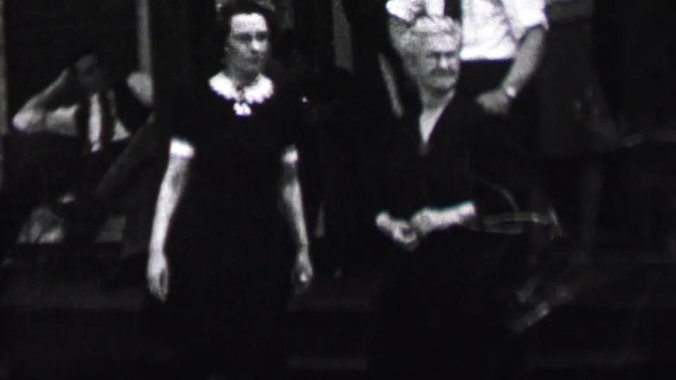 Black-and-white frame from the Albany 1937 home movie showing two women in dark dresses near outdoor steps, one looking forward and another turning to the side, with suited men behind them.