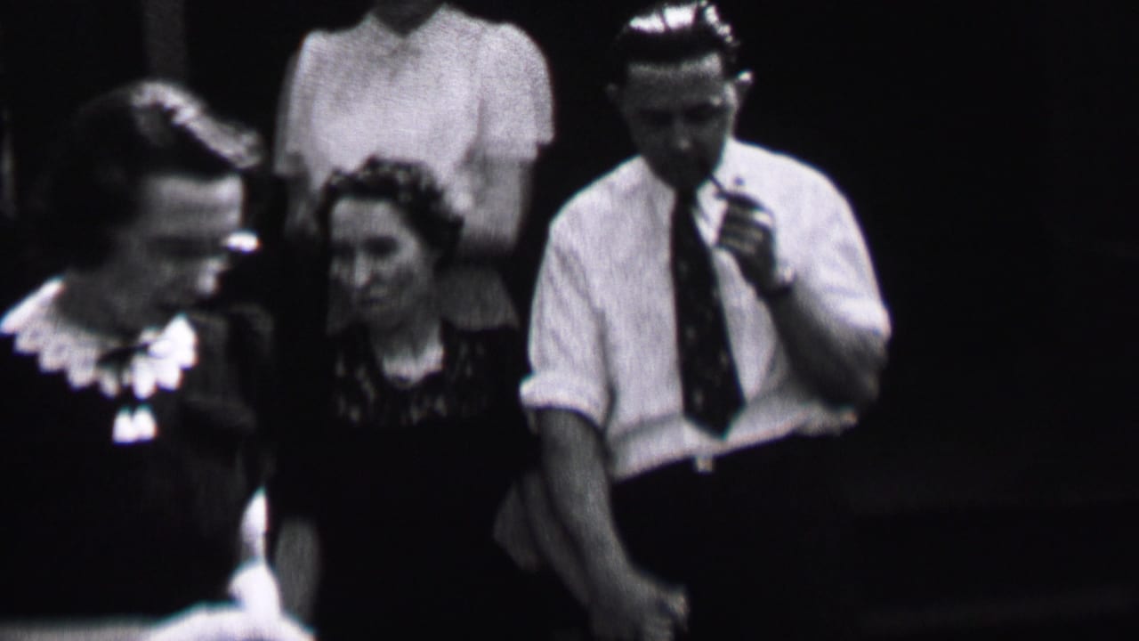 A man in a white shirt and tie smokes a pipe while women stand nearby.